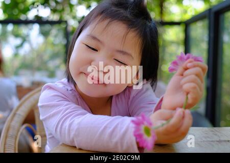 Ein nettes schüchternes asiatisches Mädchen, das an einem Tisch in einem Café sitzt, entspannt und ihre schönen rosa Blumen genießt, lächelnd. Stockfoto