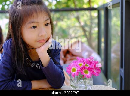 Ein süßes asiatisches Mädchen mit ihrem Gesicht, das auf ihrer Hand liegt, an einem Couchtisch mit schönen rosa Gänseblümchen sitzt, mit wunderbaren Gedanken nachdenkend, wartet Stockfoto