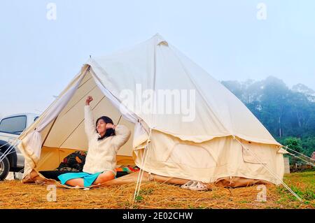 Eine junge Frau sitzt friedlich in ihrem weißen Zelt am Morgen mit kühler Brise und Morgensonne, Stretching und entspannend, zeigt in die DIS Stockfoto