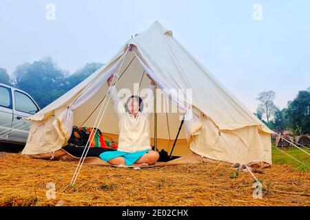 Eine junge Frau sitzt friedlich in ihrem weißen Zelt am Morgen mit kühler Brise und Morgensonne, Stretching und entspannend, zeigt in die DIS Stockfoto