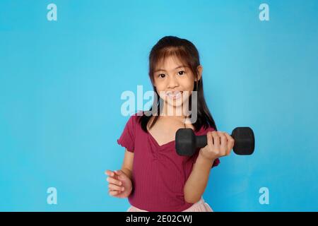 Ein süßes asiatisches Mädchen hält eine Hantel, trainieren, glücklich und stark fühlen. Einfacher hellblauer Hintergrund. Stockfoto