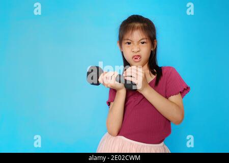 Ein süßes asiatisches Mädchen hält eine schwere Hantel, die Ausübung, macht lustige Gesichter. Einfacher hellblauer Hintergrund. Stockfoto