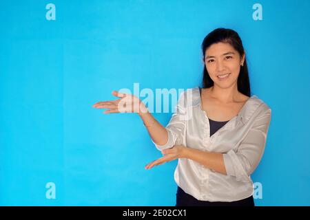 Eine Aufnahme in Taillenhöhe einer attraktiven asiatischen Frau in den 30er Jahren, die ein Produkt präsentiert und mit offenen Händen auf einen hellblauen, schlichten Hintergrund zeigt. Stockfoto