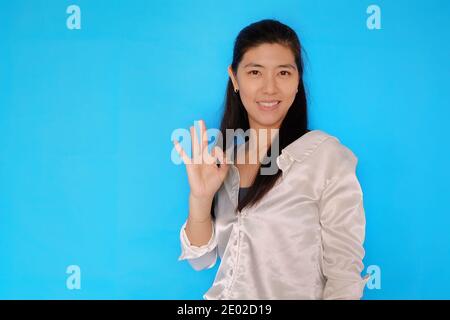 Eine attraktive junge asiatische Frau, die mit der rechten Hand ein Zeichen für Ordnung macht, um zu bestätigen, dass alles in Ordnung ist. Mit hellblauem, einfarbigem Hintergrund. Stockfoto