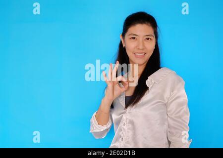 Eine attraktive junge asiatische Frau, die mit der rechten Hand ein Zeichen für Ordnung macht, um zu bestätigen, dass alles in Ordnung ist. Mit hellblauem, einfarbigem Hintergrund. Stockfoto