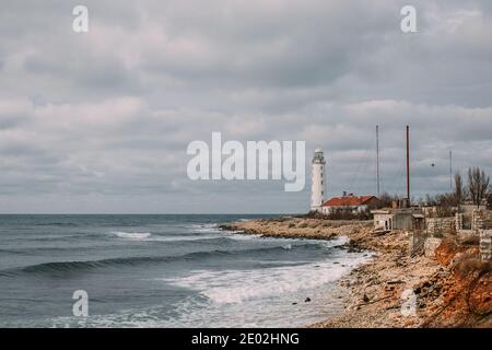 Wunderschöne Meereslandschaft mit einem weißen Leuchtturm und alten Gebäuden am Ufer. Es gibt ein welliges Meer unten, und ein wolkiger Himmel oben. Stockfoto