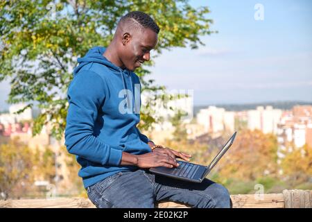 Afrikanischer schwarzer Mann lächelt und arbeitet mit einem Laptop in einem Park. Digital Nomad Entrepreneur Konzept. Stockfoto