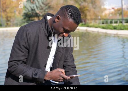 Junger afrikanischer schwarzer Mann lacht beim Chatten auf dem Smartphone in einem Park. Geschäftsmann am Telefon. Stockfoto