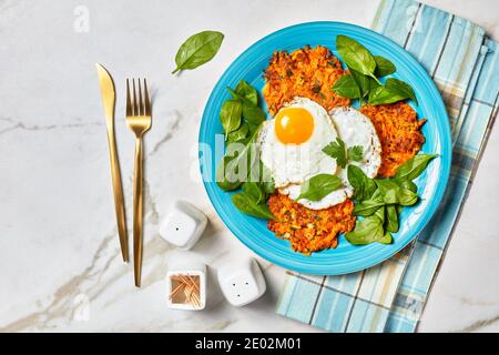 Süße Kartoffelrosti mit gebratenen Eiern und Grüns auf einem blauen Teller auf einem Marmortisch serviert, amerikanisches Frühstück am Morgen, horizontale Ansicht von oben, flach l Stockfoto