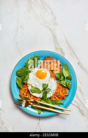 Süße Kartoffelpfannkuchen mit gebratenen Eiern und Grüns auf einem blauen Teller auf einem Marmortisch serviert, amerikanisches Frühstück am Morgen, horizontale Ansicht von oben, Fla Stockfoto