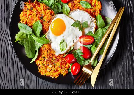 Karotten Pfannkuchen mit gebratenen Eiern und Grünpflanzen auf einem schwarzen Teller auf einem Holztisch serviert, horizontale Ansicht von oben, flach liegend, Makro Stockfoto