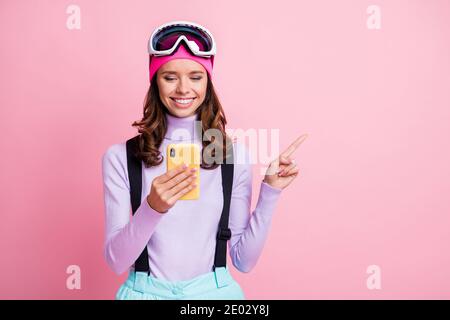 Foto-Porträt einer lächelnden Frau, die mit dem Finger auf einen leeren Raum zeigt Halten Telefon in einer Hand isoliert auf pastellrosa gefärbt Hintergrund Stockfoto