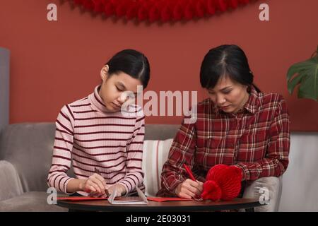 Mittleres Porträt der modernen chinesischen Frau und ihrer Tochter sitzend Auf dem Sofa machen rote Umschläge für Lunar Neujahr Geschenke Stockfoto