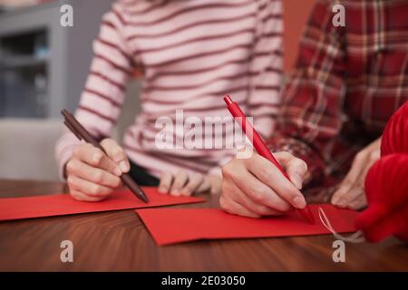 Unerkennbare chinesische Frau und Mädchen sitzen zusammen am Holztisch Dekorieren von handgefertigten roten Umschlägen für Geldgeschenke Stockfoto