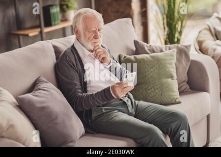 Seitenfoto von schönen schweren alten Mann gekleidet weißen Hemd Halten moderne Gerät Sofa im Inneren Wohnung Haus sitzen Stockfoto