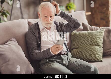 Foto von niedlichen nachdenklich Rentner tragen graue Strickjacke Sitzcouch Halten moderne Gadget drinnen Haus Wohnung Stockfoto