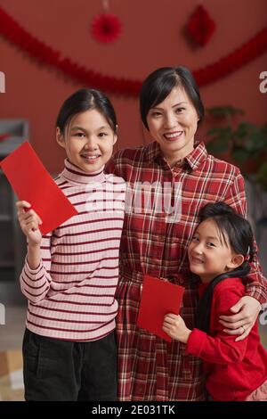 Vertikale mittel lange Schuss freudige reife chinesische Frau stehen Mit ihren schönen Töchtern, die rote Umschläge halten Stockfoto