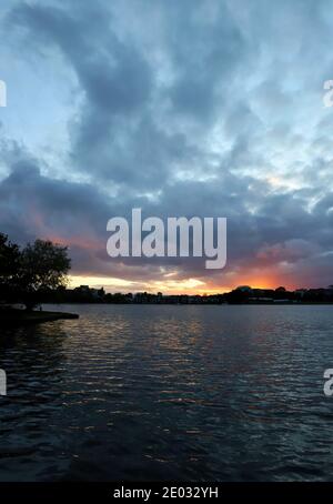 Regenwolken füllen den Himmel an einem unruhigen Sommerabend, wenn die Sonne in einer orangen Farbe über dem plätschernden Wasser des Poole Park untergeht. Stockfoto