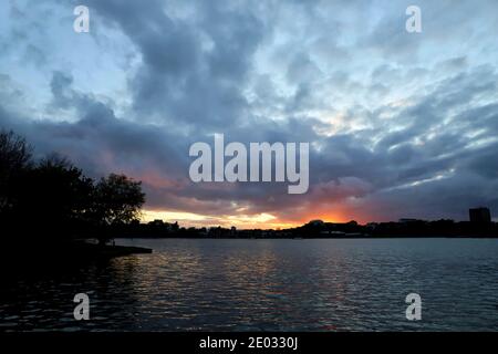 Regenwolken füllen den Himmel an einem unruhigen Sommerabend, wenn die Sonne in einer orangen Farbe über dem plätschernden Wasser des Poole Park untergeht. Stockfoto