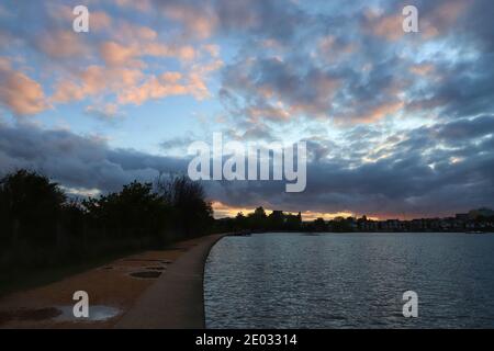 Regenwolken füllen den Himmel an einem unruhigen Sommerabend, wenn die Sonne in einer orangen Farbe über dem plätschernden Wasser des Poole Park untergeht. Stockfoto