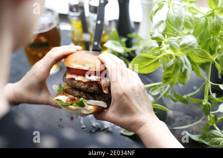 Hamburger mit zwei Rinderschnitzel, Tomate, Salat und Sauce. Burger auf einem Teller. Frau isst einen Hamburger. Appetitliches Gericht auf einem schwarzen Teller serviert Stockfoto