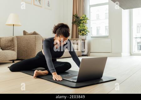 Junge Frau aus dem Nahen Osten, die zu Hause Yoga macht. Frau in Sportkleidung sitzt auf einer Matte und streckt ihre Beine vor einem Laptop. Stockfoto