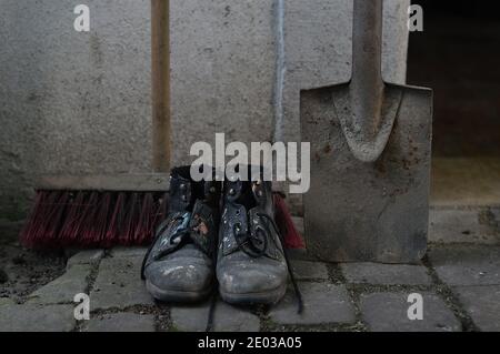 Alte abgenutzte, lackierte Arbeitsstiefel mit einem Hofbeer und einem Spaten als Symbol für Reinigung, Hausmeister oder Wartung Stockfoto