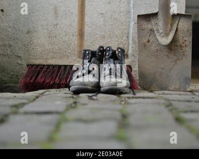 Alte abgenutzte, lackierte Arbeitsstiefel mit einem Hofbeer und einem Spaten als Symbol für Reinigung, Hausmeister oder Wartung Stockfoto