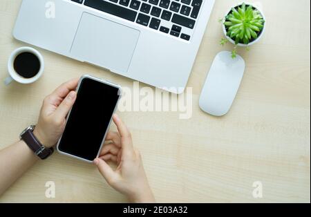 Hand mit Uhr, die Smartphone auf einem Holzschreibtisch mit Computer, Maus, Topfpflanze und Kaffee hält. Speicherplatz kopieren Stockfoto