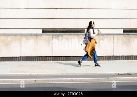 London, Großbritannien, 11. September 2020: Eine junge Frau, die mit ihrem Kaffee mit Gesichtsmaske durch leere Londoner Straßen läuft Stockfoto