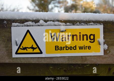 Gelb, schwarz und weiß rechteckiges Schild: Warnung Stacheldraht mit Stacheldraht-Symbol in Dreieck. Auf Holzzaun mit Schnee auf dem Zaun. Stockfoto