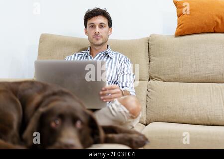 Mann, der mit einem Laptop auf dem Sofa arbeitet, während er zugewandt ist Die Kamera mit einem labrador-Hund, der neben ihm liegt Stockfoto