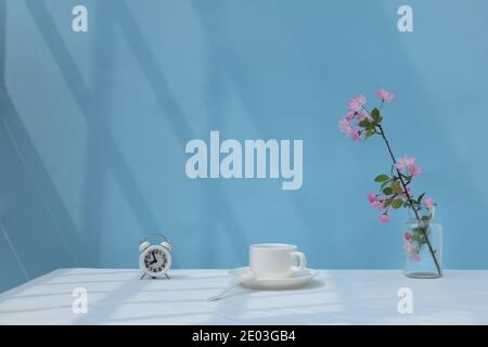 Pfirsichblüten, eine Tasse Kaffee, Wecker auf dem Schreibtisch, Sonnenlicht durch das Fenster auf dem Schreibtisch des Heimbüros, minimalistisch Stockfoto