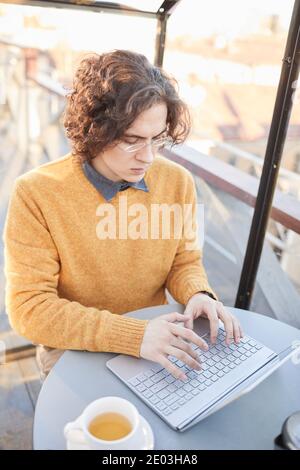 Junger Mann mit lockigen Haaren in Brillen sitzen an der Tisch im Café und Online-Arbeit auf Laptop Stockfoto