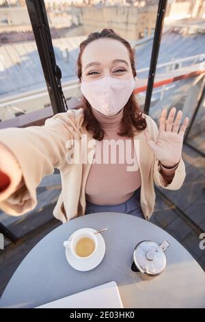 Selfie-Porträt einer jungen Frau in Schutzmaske winkt an Kamera auf ihrem Handy im Café Stockfoto