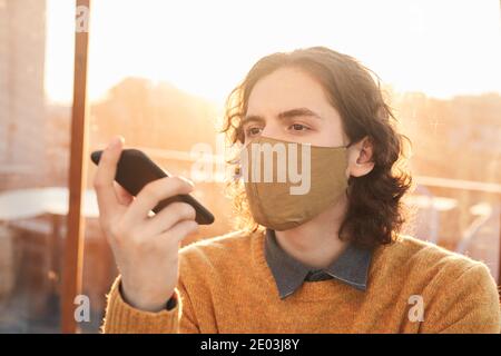 Junger Mann in Schutzmaske hört auf Audio-Nachricht auf Sein Mobiltelefon Stockfoto
