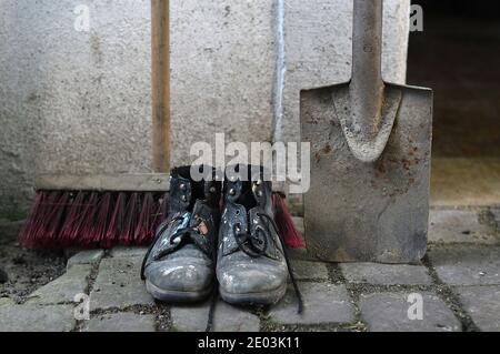 Alte abgenutzte, lackierte Arbeitsstiefel mit einem Hofbeer und einem Spaten als Symbol für Reinigung, Hausmeister oder Wartung Stockfoto