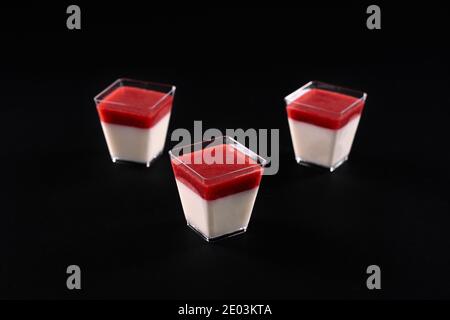 Vorderansicht von drei kleinen quadratischen Gläsern mit milchiger Panna Cotta und Erdbeermarmelade. Süßes köstliches Dessert mit leuchtend roten Belag dekoriert, isoliert auf schwarzem Hintergrund. Food-Konzept. Stockfoto