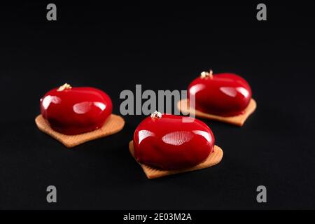 Nahaufnahme von roten Kuchen mit Gold isoliert auf schwarzem Hintergrund dekoriert. Dessert mit glatter Oberfläche, Mousse und Spiegelglasur auf süßen Plätzchen, Herzform. Köstliches Gericht in der Cafeteria. Stockfoto