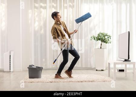 Fröhlicher Mann, der mit einem Putzmopp singt und tanzt Zuhause vor dem fernseher Stockfoto