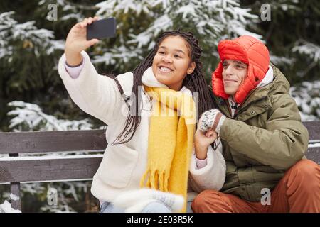 Junge glückliche Frau macht Selfie auf ihrem Handy mit Ihr Freund während sie im Winter auf der Bank sitzen Stockfoto