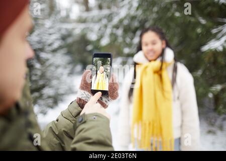 Nahaufnahme eines jungen Mannes, der das Mobiltelefon hält und das fotografiert Frau, die sich gegen die verschneiten Bäume im Wald posiert Stockfoto