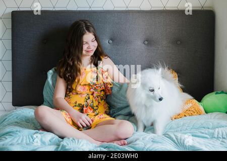 Junges Mädchen sitzt auf einem Bett patting eine niedliche kleine Flauschiger weißer Hund Stockfoto