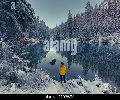 See Prags Italienische Dolomitenperson von hinten mit gelber Jacke Am Ufer eines Bergsees mit Blick auf die Blick auf verschneite Bäume Stockfoto