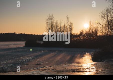 Orange lebhafter Sonnenuntergang auf eisig gefrorenen reflektierenden See mit dunklen Bäumen Silhouetten, langen Schatten und klaren Himmel Stockfoto