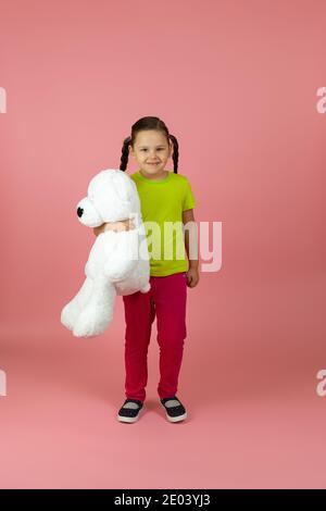 Ein durchgehendes zufriedenes, glückliches pigtailed Mädchen in einem hellgrünen T-Shirt und roter Hose hält einen weißen Teddybär isoliert auf einem rosa Hintergrund Stockfoto