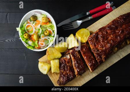 Rippchen in Grillsauce und Kartoffeln auf einem Holzbrett mit schwarzem Hintergrund. Stockfoto