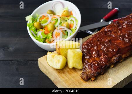 Grillrippen mit Kartoffeln und Salat auf einem Holzbrett auf dunklem Hintergrund. Speicherplatz kopieren. Stockfoto