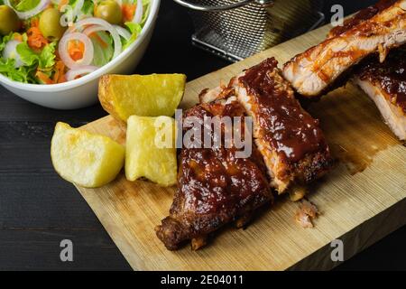 Grillrippen mit Kartoffeln und Salat auf einem Holzbrett auf dunklem Hintergrund. Stockfoto