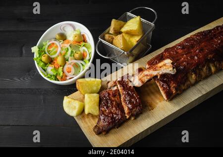 Grillrippen mit Kartoffeln und Salat auf einem Holzbrett auf dunklem Hintergrund. Speicherplatz kopieren. Stockfoto